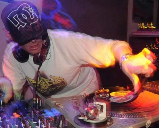 DJ Natural Nate® in DC hat and BYBB white tee, motion blur scratch action under warm orange stage light
